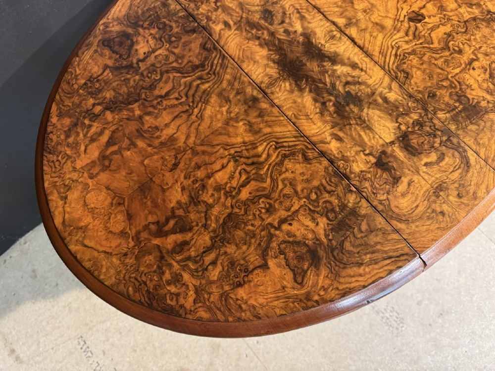 An antique Victorian burr walnut Sutherland table featuring an oval drop-leaf top, turned support legs, and small castors. View VII