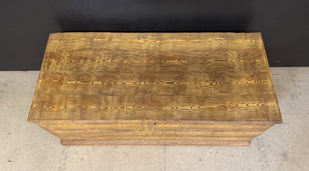 A low rectangular antique pine chest featuring prominent wood graining, a hinged flat lid, central keyhole, and a solid plinth base. View II