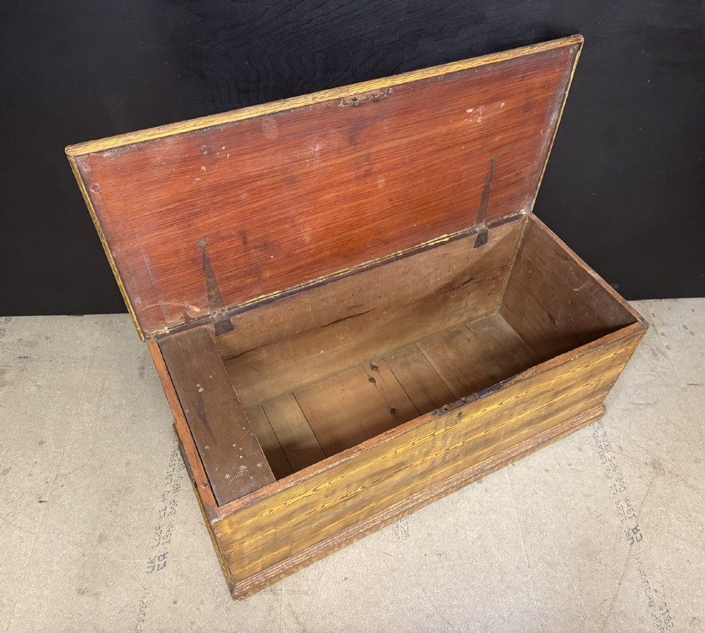A low rectangular antique pine chest featuring prominent wood graining, a hinged flat lid, central keyhole, and a solid plinth base. View V