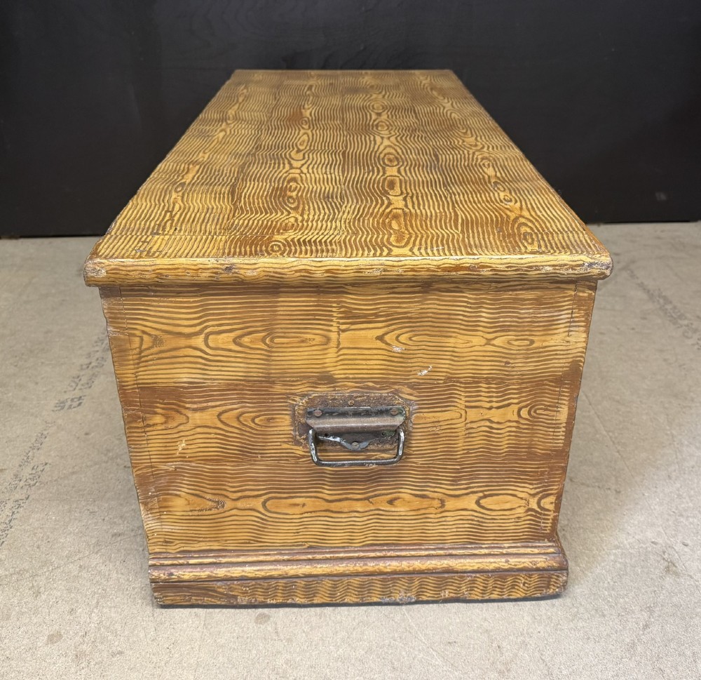 A low rectangular antique pine chest featuring prominent wood graining, a hinged flat lid, central keyhole, and a solid plinth base. View VII