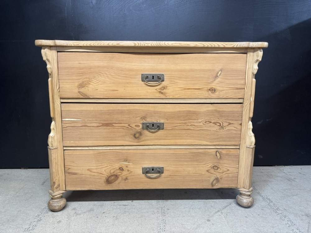 A three-drawer stripped pine chest of drawers featuring carved side corbels, brass drop handles, and turned bun feet. View II