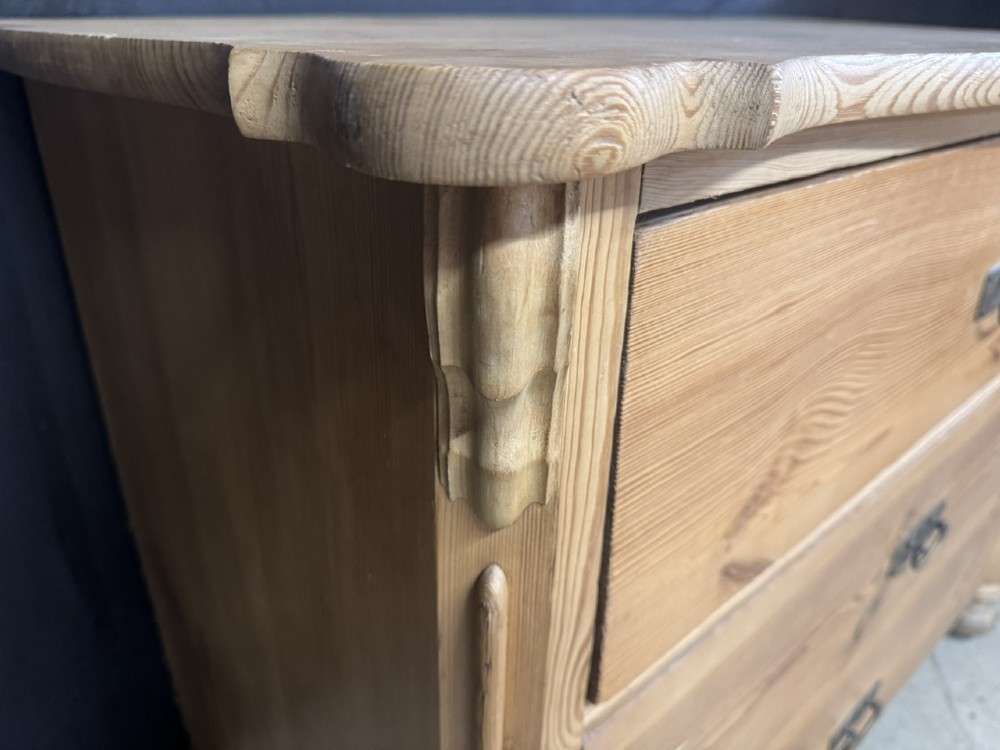 A three-drawer stripped pine chest of drawers featuring carved side corbels, brass drop handles, and turned bun feet. View VI