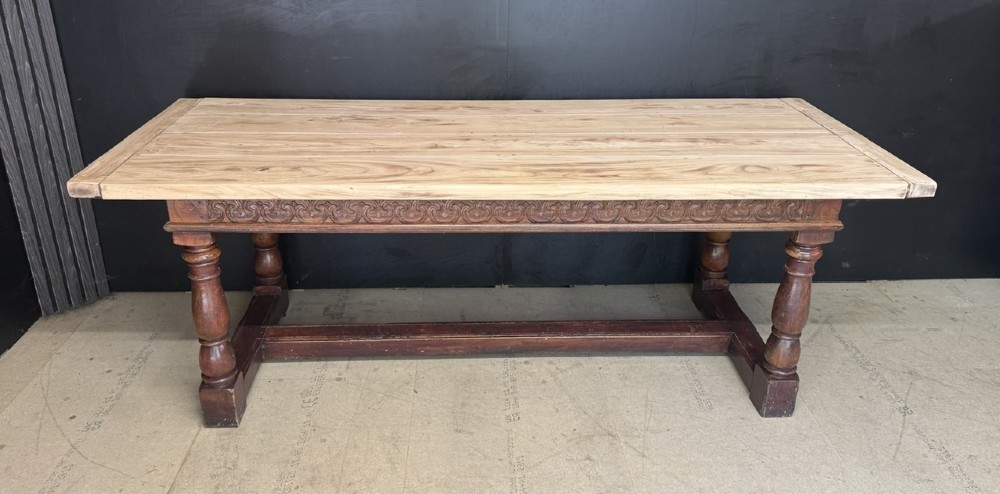 A rustic wooden dining table with a natural scrubbed timber top, carved apron frieze, turned legs, and a floor-level H-stretcher.