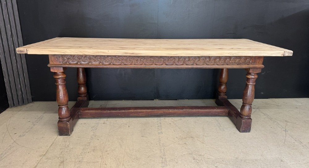 A rustic wooden dining table with a natural scrubbed timber top, carved apron frieze, turned legs, and a floor-level H-stretcher. View II