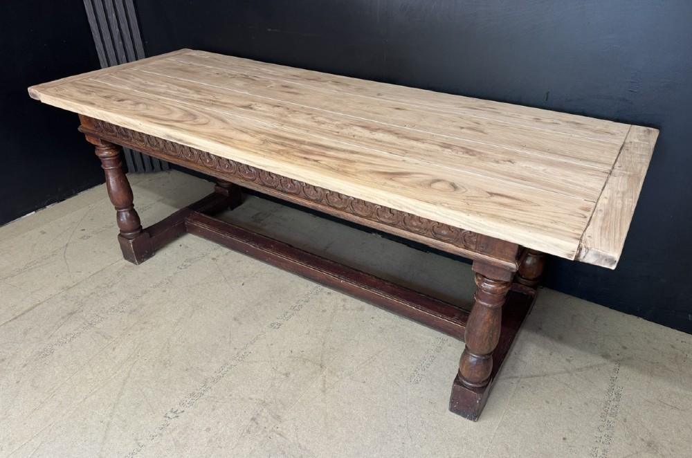 A rustic wooden dining table with a natural scrubbed timber top, carved apron frieze, turned legs, and a floor-level H-stretcher. View III