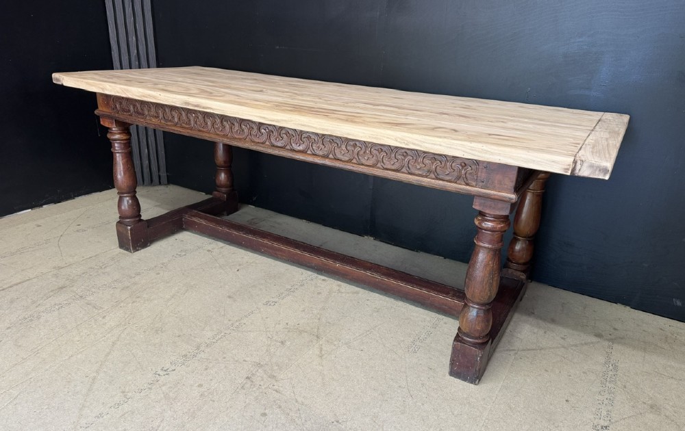 A rustic wooden dining table with a natural scrubbed timber top, carved apron frieze, turned legs, and a floor-level H-stretcher. View IV
