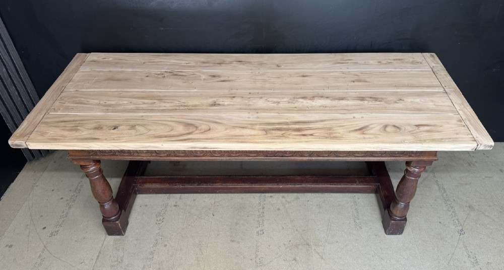 A rustic wooden dining table with a natural scrubbed timber top, carved apron frieze, turned legs, and a floor-level H-stretcher. View VII