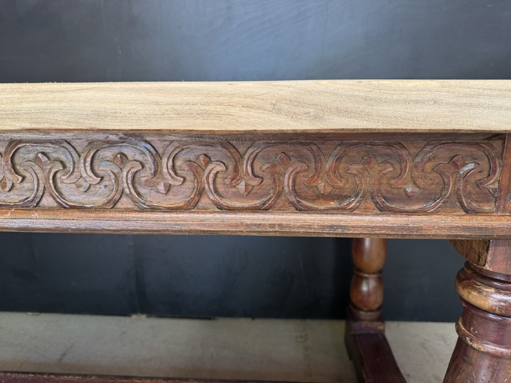 A rustic wooden dining table with a natural scrubbed timber top, carved apron frieze, turned legs, and a floor-level H-stretcher. View IX