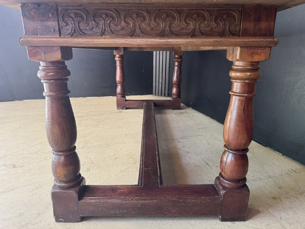 A rustic wooden dining table with a natural scrubbed timber top, carved apron frieze, turned legs, and a floor-level H-stretcher. View X