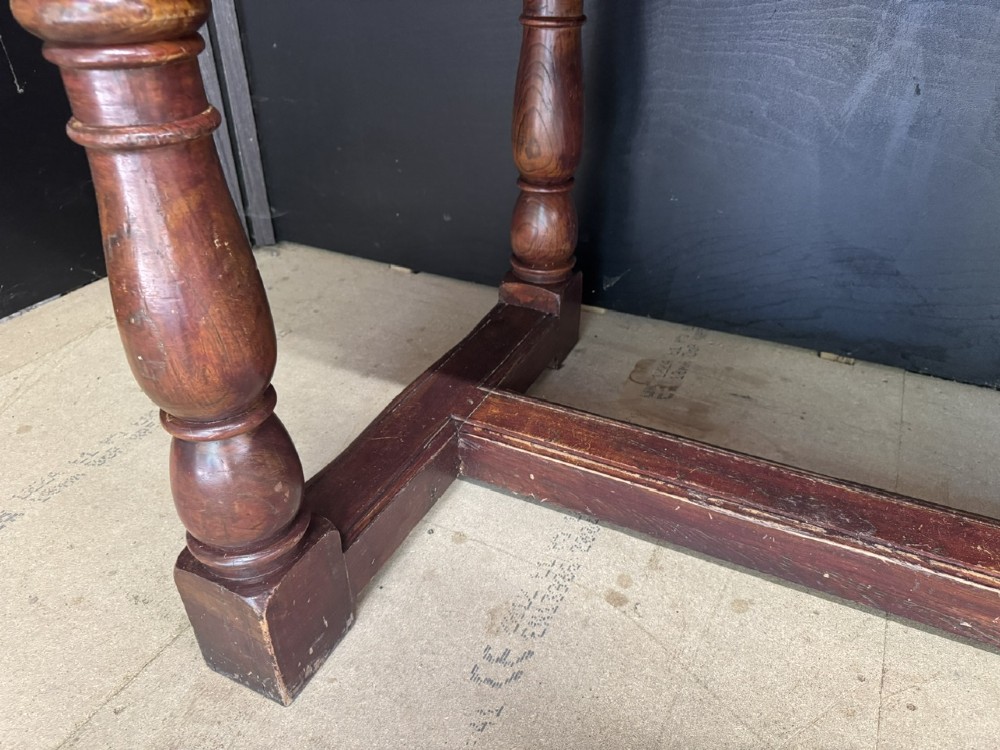 A rustic wooden dining table with a natural scrubbed timber top, carved apron frieze, turned legs, and a floor-level H-stretcher. View XII