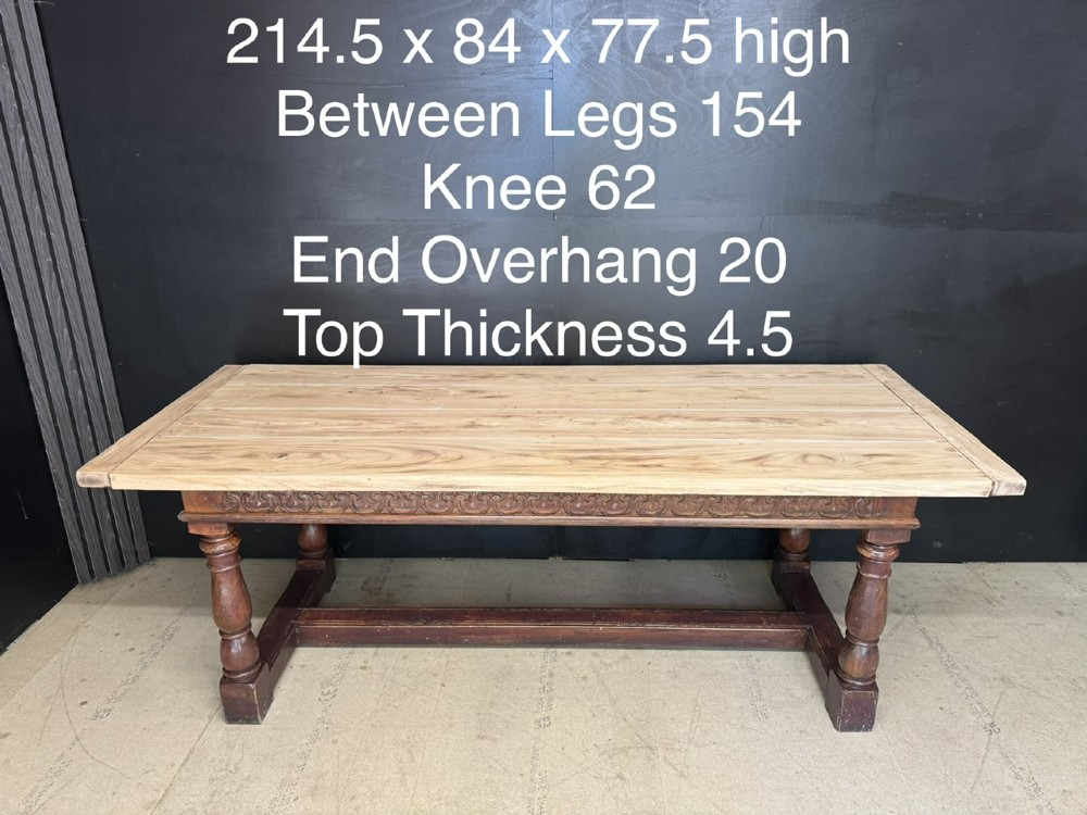 A rustic wooden dining table with a natural scrubbed timber top, carved apron frieze, turned legs, and a floor-level H-stretcher. View XV