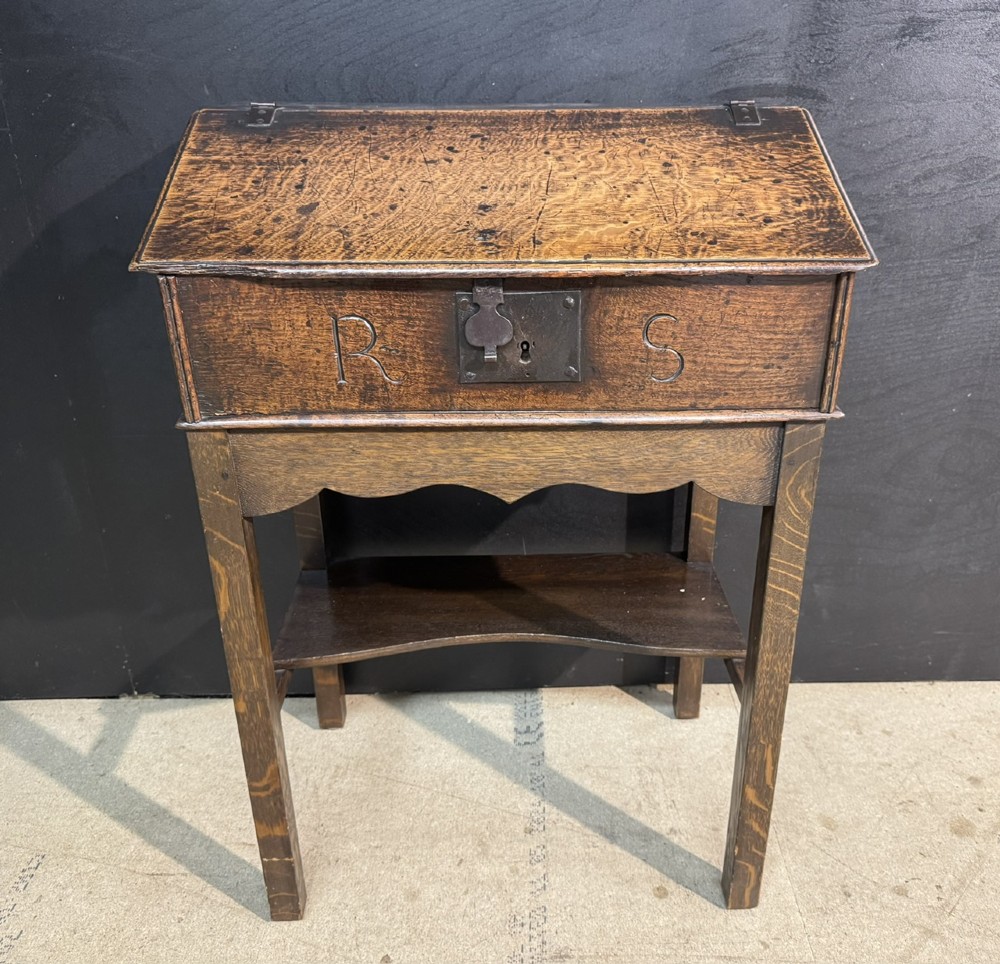 A 17th-century dark oak writing slope box on a wooden stand, featuring carved initials R S and a central iron lock plate.