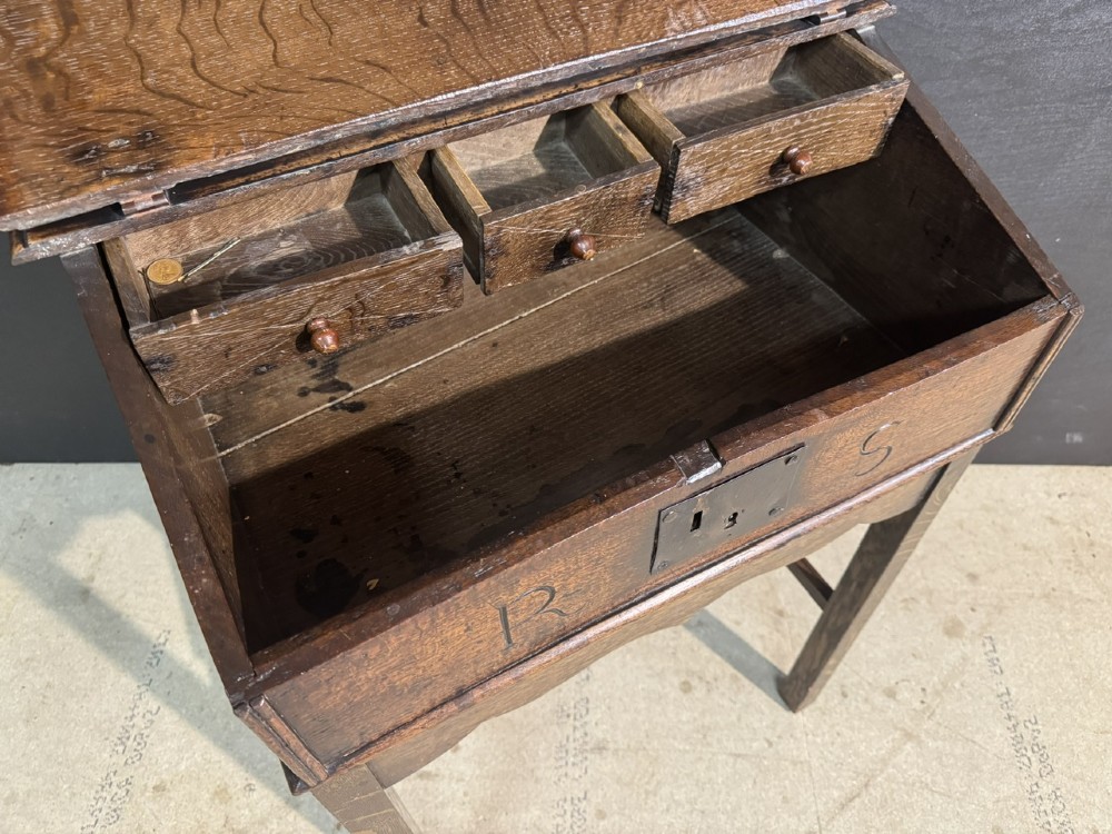 A 17th-century dark oak writing slope box on a wooden stand, featuring carved initials R S and a central iron lock plate. View VI