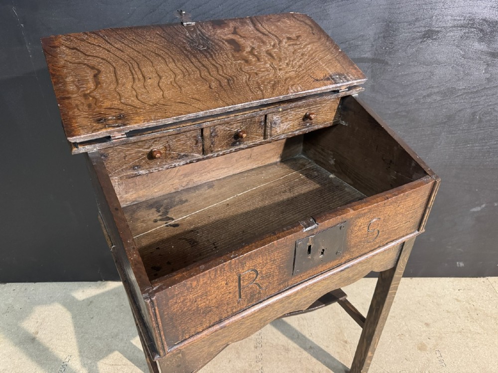 A 17th-century dark oak writing slope box on a wooden stand, featuring carved initials R S and a central iron lock plate. View VII
