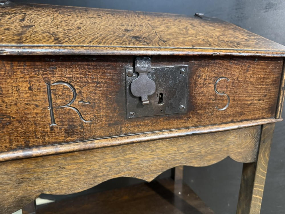 A 17th-century dark oak writing slope box on a wooden stand, featuring carved initials R S and a central iron lock plate. View VIII