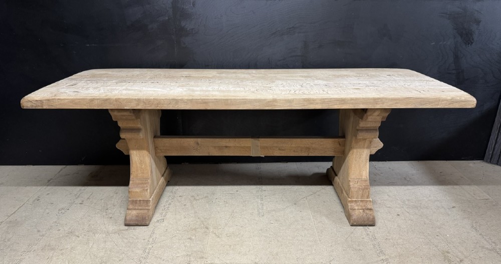 A rectangular bleached oak farmhouse dining table featuring a thick timber top supported by carved trestle legs and a central stretcher.