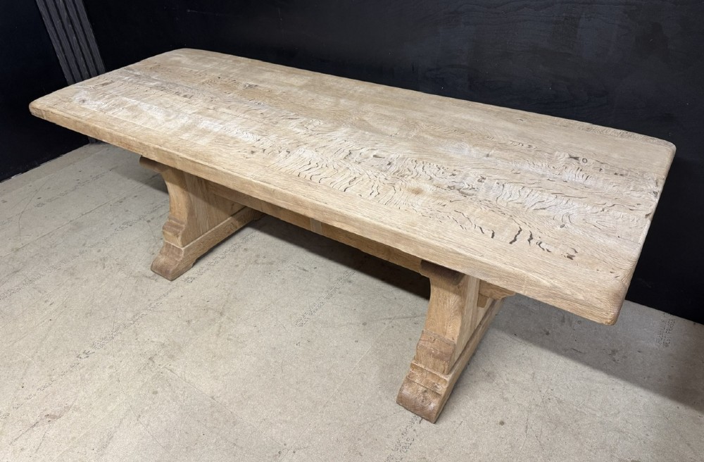 A rectangular bleached oak farmhouse dining table featuring a thick timber top supported by carved trestle legs and a central stretcher. View V