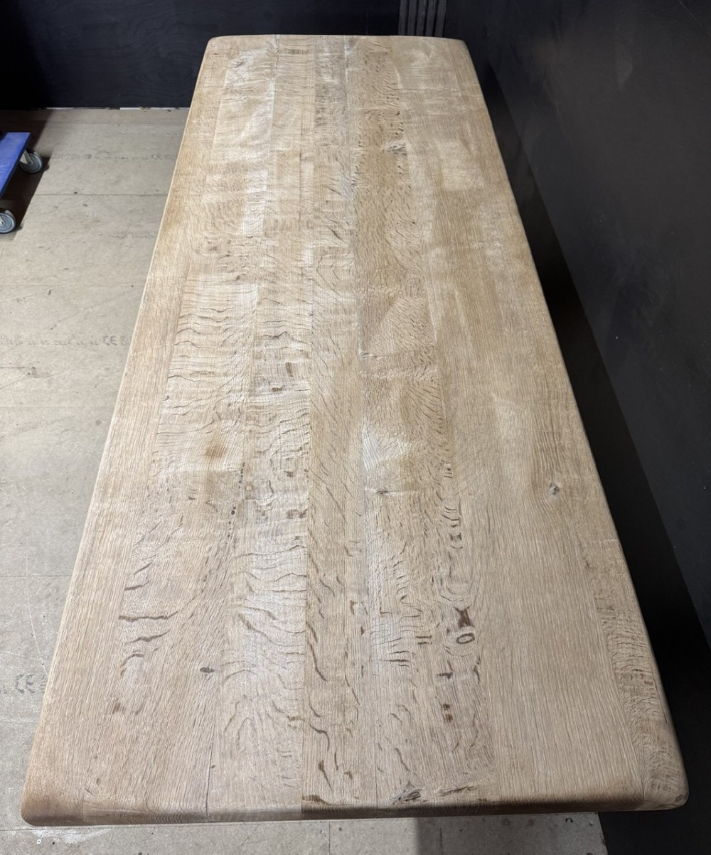 A rectangular bleached oak farmhouse dining table featuring a thick timber top supported by carved trestle legs and a central stretcher. View VI