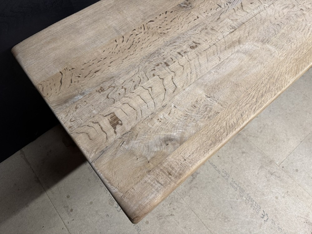 A rectangular bleached oak farmhouse dining table featuring a thick timber top supported by carved trestle legs and a central stretcher. View VII