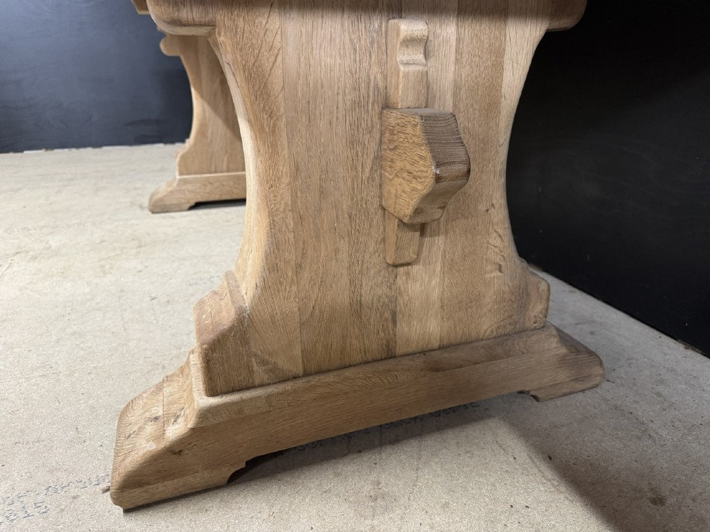 A rectangular bleached oak farmhouse dining table featuring a thick timber top supported by carved trestle legs and a central stretcher. View X