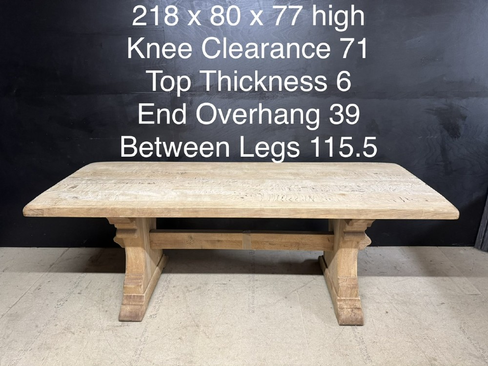 A rectangular bleached oak farmhouse dining table featuring a thick timber top supported by carved trestle legs and a central stretcher. View XII