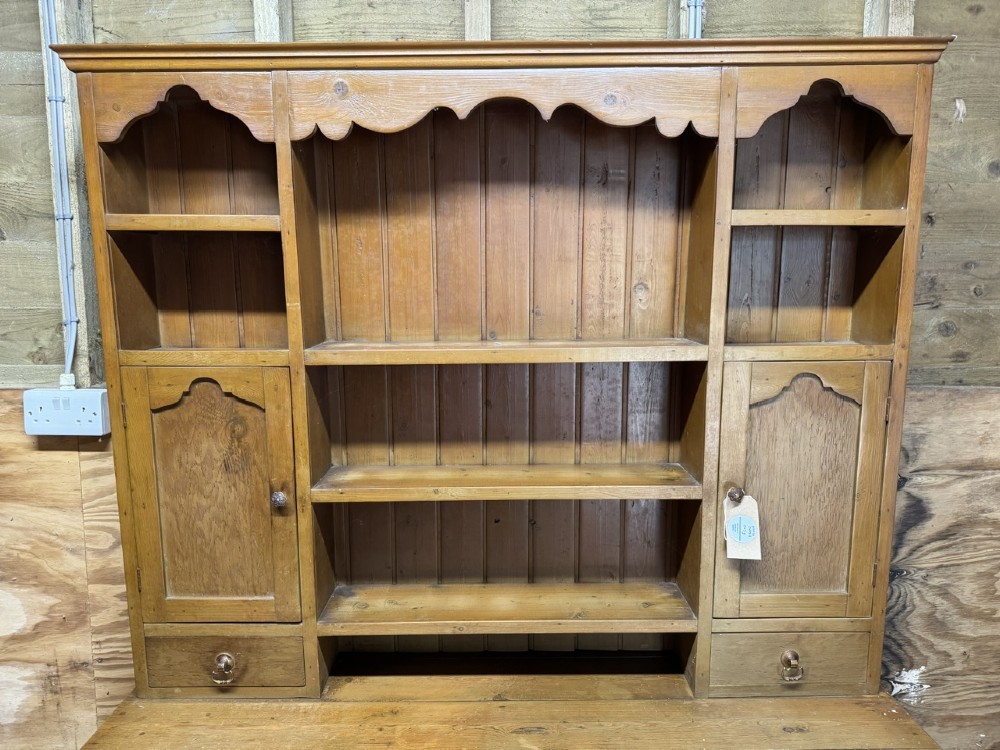 A two-part antique pine Welsh dresser featuring an open plate rack flanked by small side cupboards above three frieze drawers and an open pot board base. View V