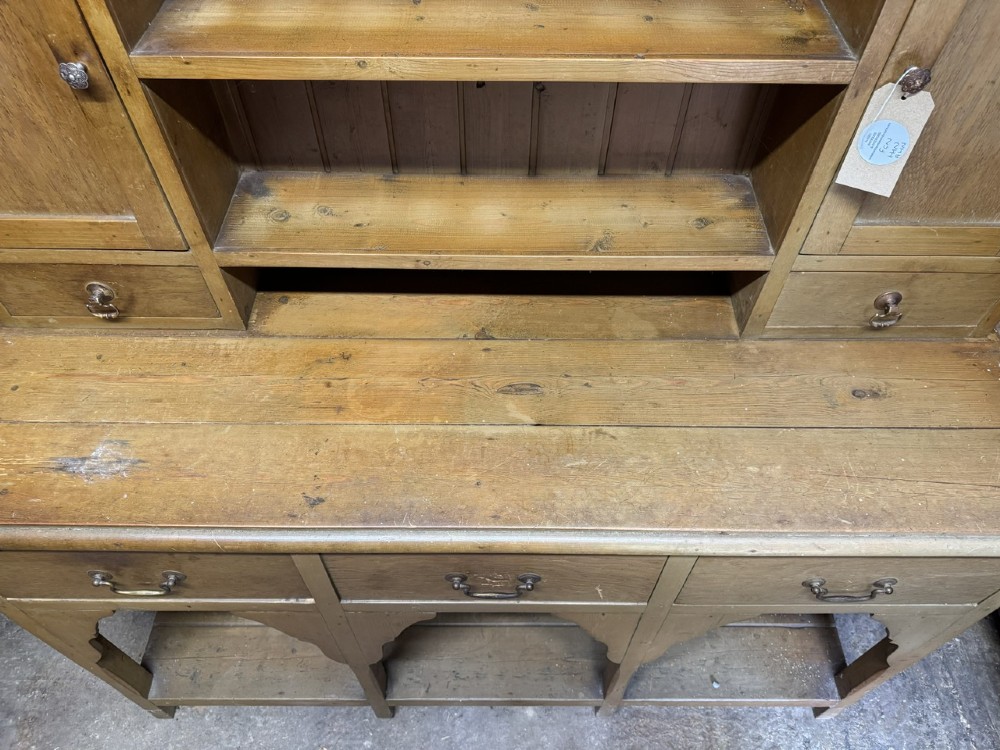 A two-part antique pine Welsh dresser featuring an open plate rack flanked by small side cupboards above three frieze drawers and an open pot board base. View X