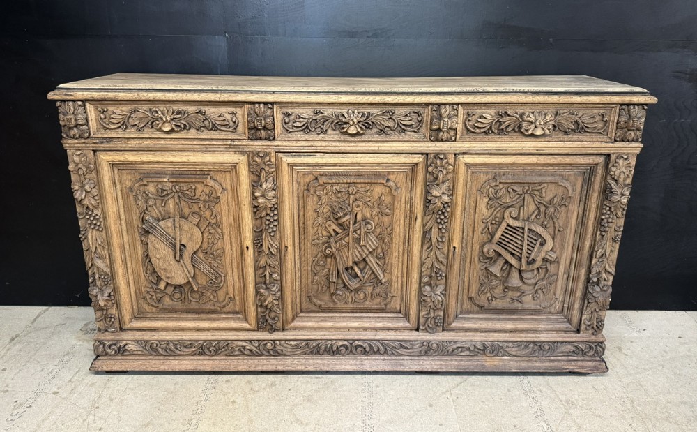 A three-door carved light oak sideboard featuring detailed relief panels depicting artistic and musical instruments.