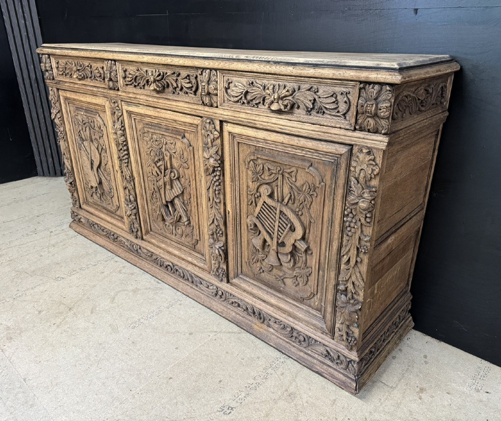 A three-door carved light oak sideboard featuring detailed relief panels depicting artistic and musical instruments. View III