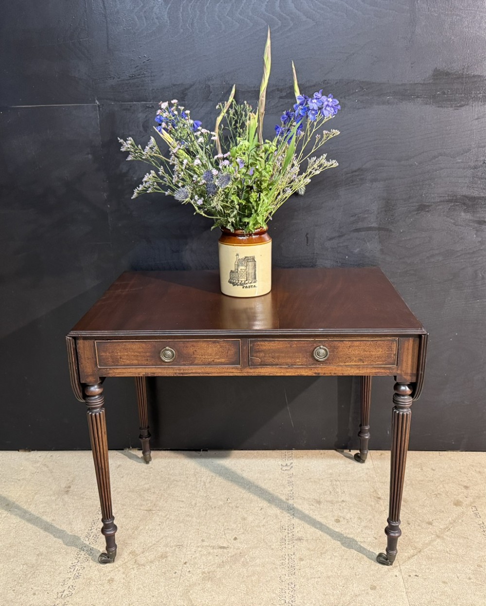 A Georgian style dark mahogany sofa table featuring two drawers with brass ring pulls, drop leaves on either side, and reeded tapered legs on brass castors. View II