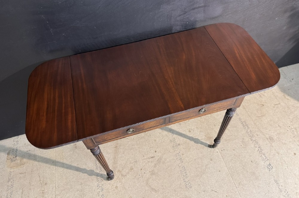 A Georgian style dark mahogany sofa table featuring two drawers with brass ring pulls, drop leaves on either side, and reeded tapered legs on brass castors. View X