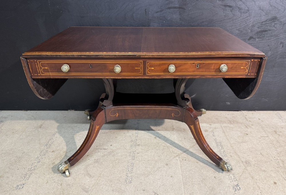 A Georgian mahogany sofa table featuring two inline drawers with satinwood string inlay, drop leaves, twin-pillar supports, and splayed legs ending in brass claw feet on castors.