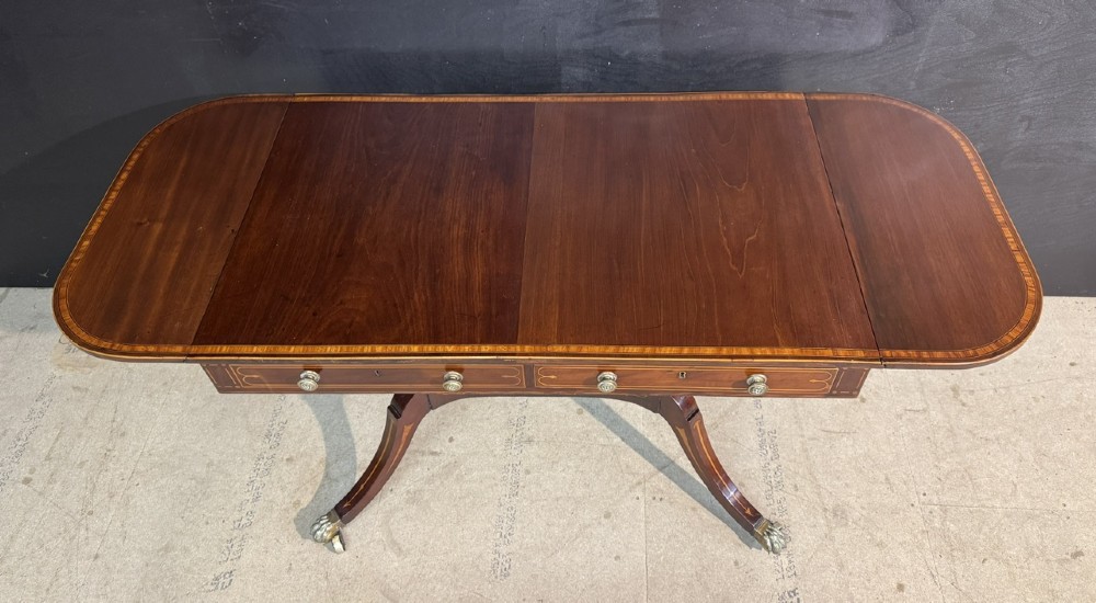 A Georgian mahogany sofa table featuring two inline drawers with satinwood string inlay, drop leaves, twin-pillar supports, and splayed legs ending in brass claw feet on castors. View V