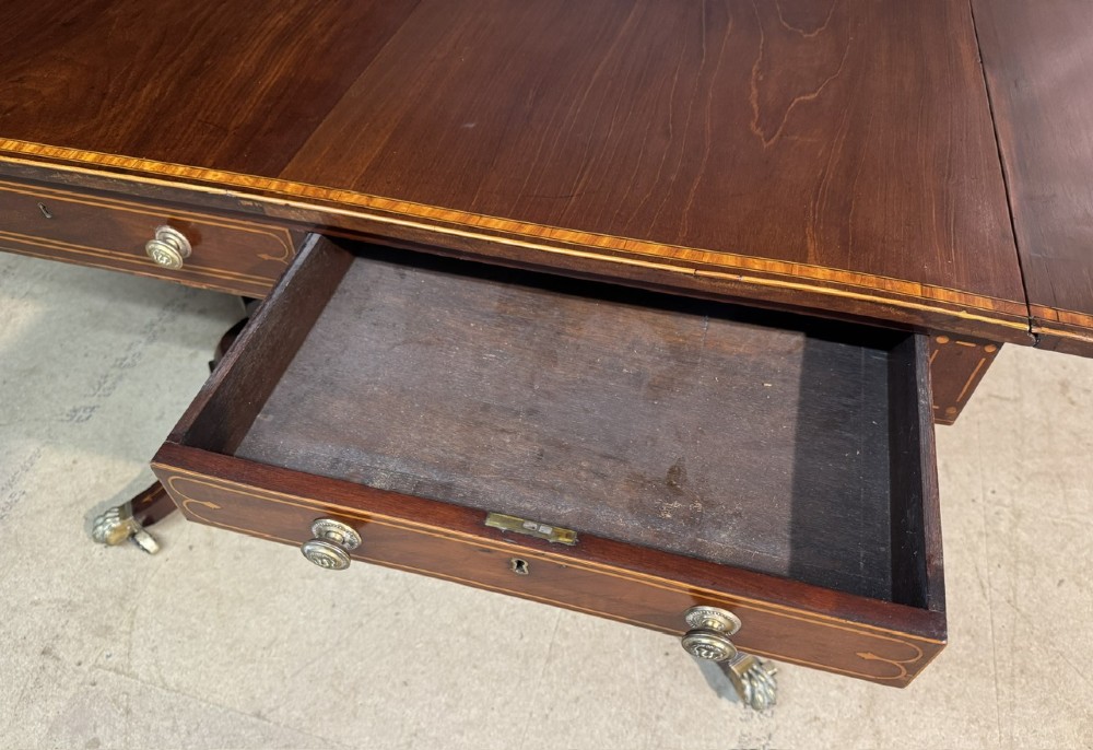 A Georgian mahogany sofa table featuring two inline drawers with satinwood string inlay, drop leaves, twin-pillar supports, and splayed legs ending in brass claw feet on castors. View X