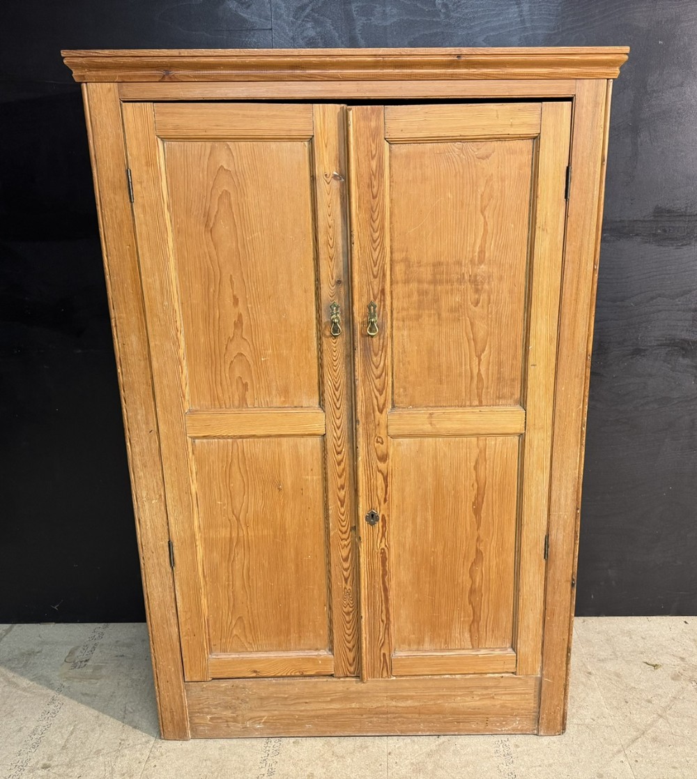 A freestanding double-door antique pine larder cupboard with four raised door panels, brass drop handles, a keyhole, and a moulded top cornice.