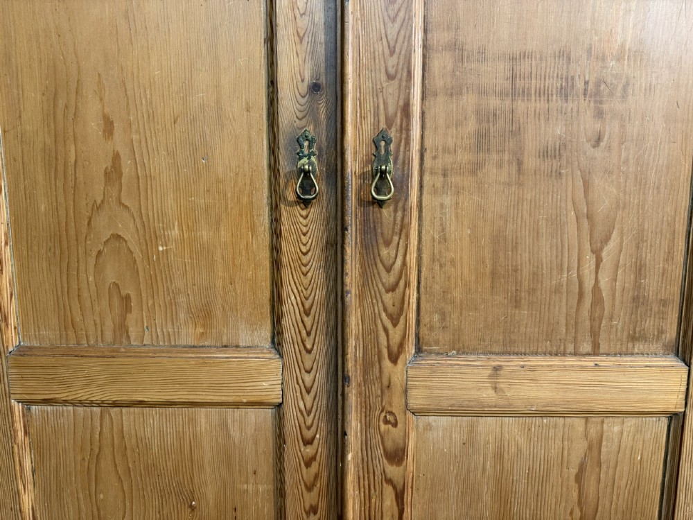 A freestanding double-door antique pine larder cupboard with four raised door panels, brass drop handles, a keyhole, and a moulded top cornice. View II
