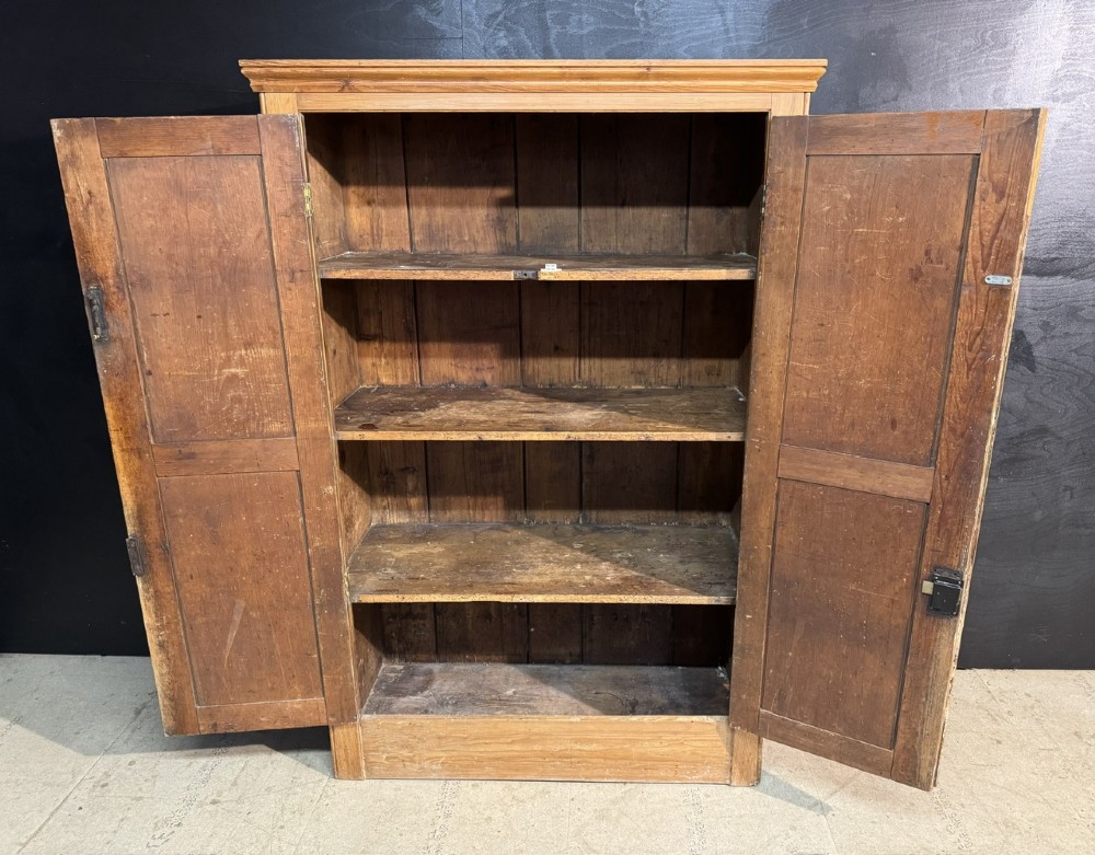 A freestanding double-door antique pine larder cupboard with four raised door panels, brass drop handles, a keyhole, and a moulded top cornice. View IV
