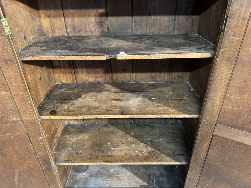 A freestanding double-door antique pine larder cupboard with four raised door panels, brass drop handles, a keyhole, and a moulded top cornice. View V