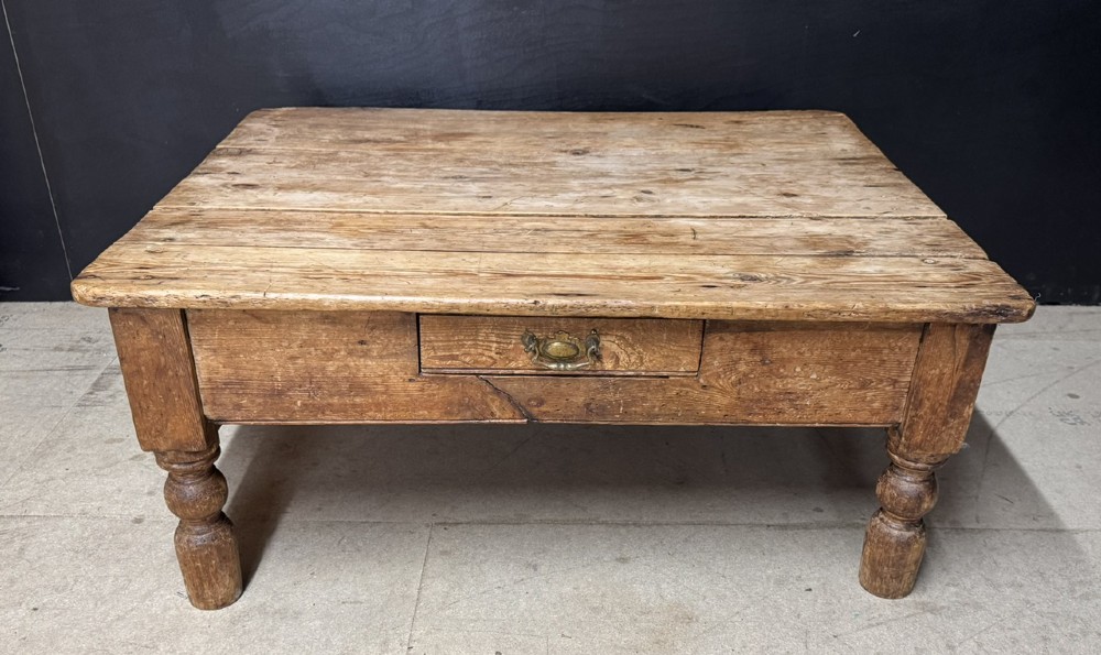 A low rectangular rustic pine coffee table featuring a plank top, turned corner legs, and a single central drawer with a brass drop handle.