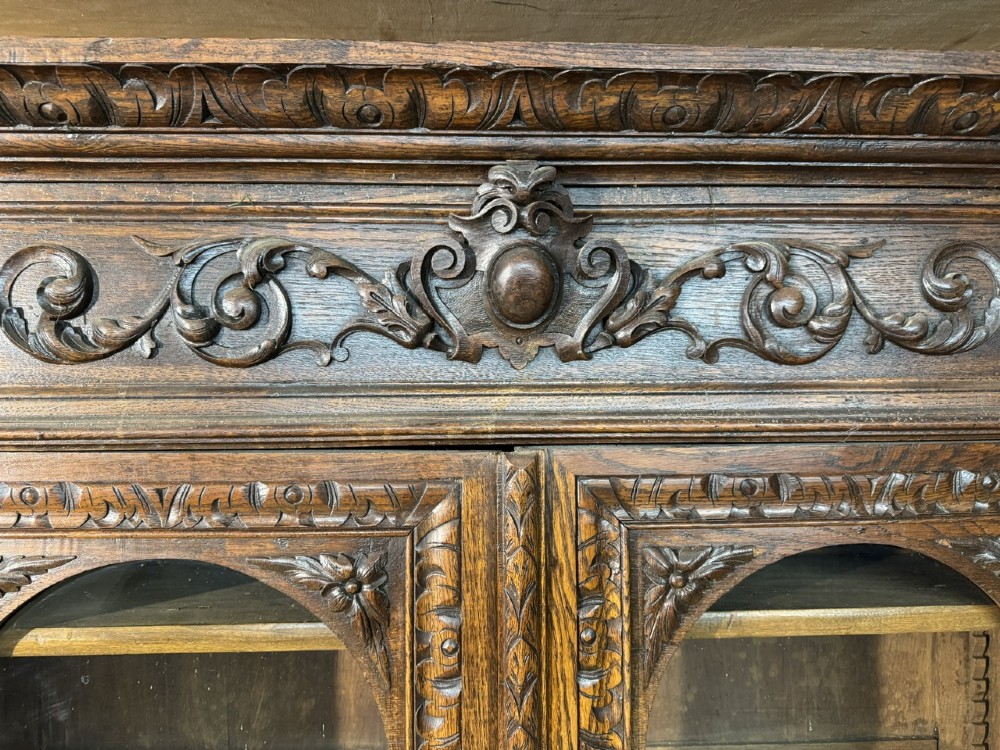 A two-tier dark oak cabinet featuring arched glazed top doors, carved scroll motifs, and lower doors with relief carvings of game birds. View VII