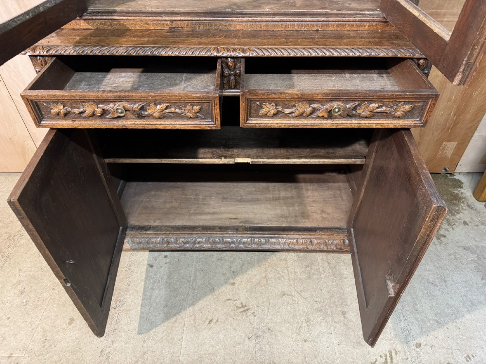 A two-tier dark oak cabinet featuring arched glazed top doors, carved scroll motifs, and lower doors with relief carvings of game birds. View VIII