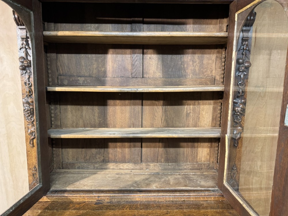 A two-tier dark oak cabinet featuring arched glazed top doors, carved scroll motifs, and lower doors with relief carvings of game birds. View IX