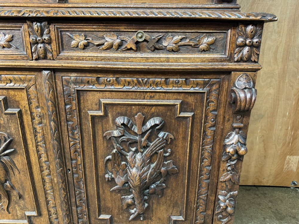 A two-tier dark oak cabinet featuring arched glazed top doors, carved scroll motifs, and lower doors with relief carvings of game birds. View XII