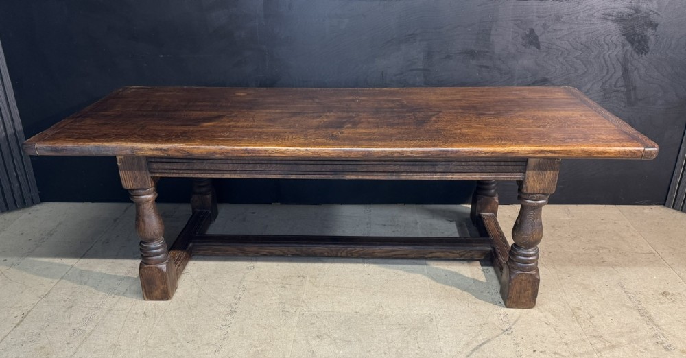 A long and deep solid dark oak farmhouse dining table featuring a plank top, moulded apron, and thick turned baluster legs joined by a floor-level stretcher.