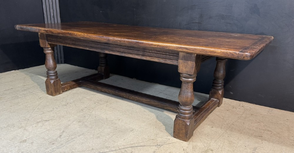 A long and deep solid dark oak farmhouse dining table featuring a plank top, moulded apron, and thick turned baluster legs joined by a floor-level stretcher. View III