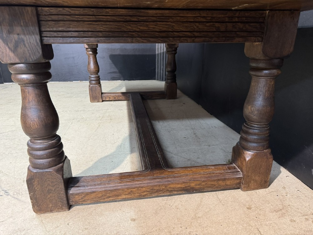 A long and deep solid dark oak farmhouse dining table featuring a plank top, moulded apron, and thick turned baluster legs joined by a floor-level stretcher. View IX
