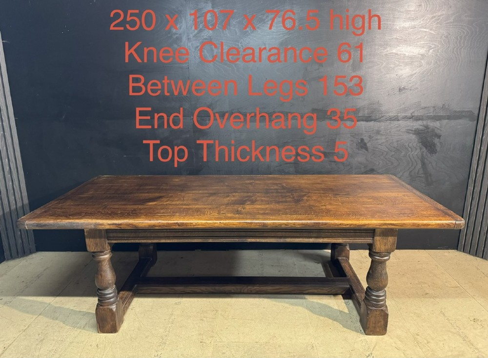 A long and deep solid dark oak farmhouse dining table featuring a plank top, moulded apron, and thick turned baluster legs joined by a floor-level stretcher. View XV