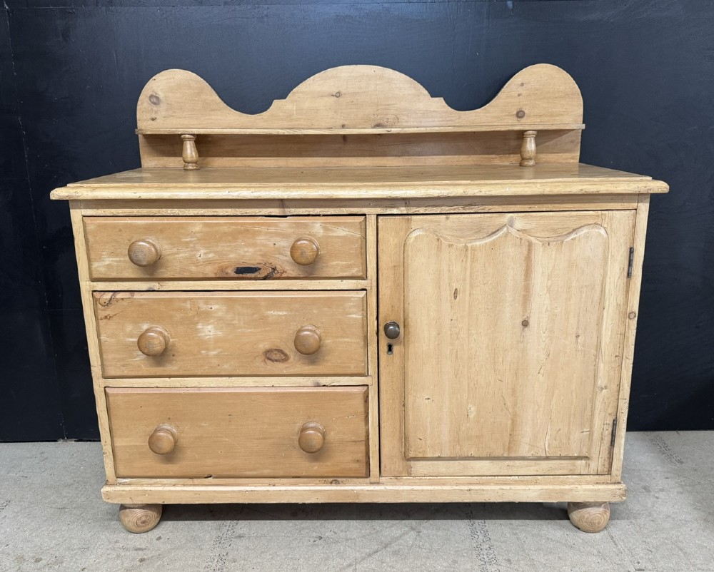 A small antique pine dresser base featuring three drawers on the left, a single panelled cupboard on the right, wooden knobs, turned bun feet, and a shaped back gallery shelf. View II