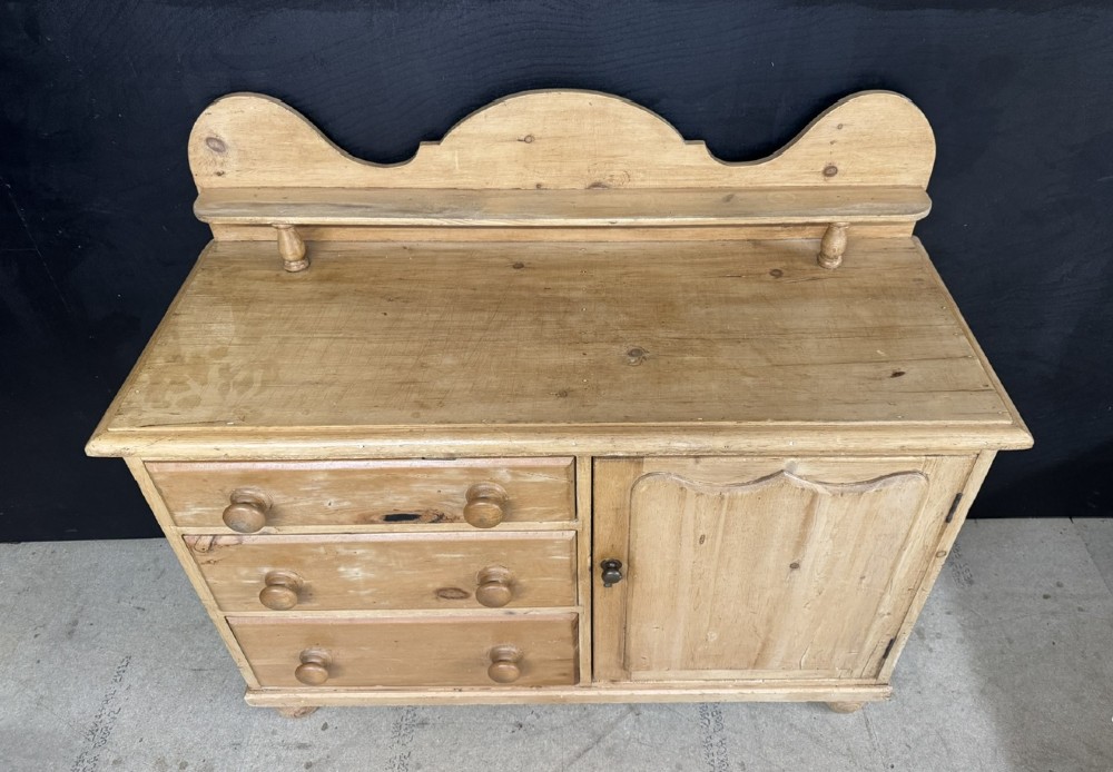 A small antique pine dresser base featuring three drawers on the left, a single panelled cupboard on the right, wooden knobs, turned bun feet, and a shaped back gallery shelf. View III