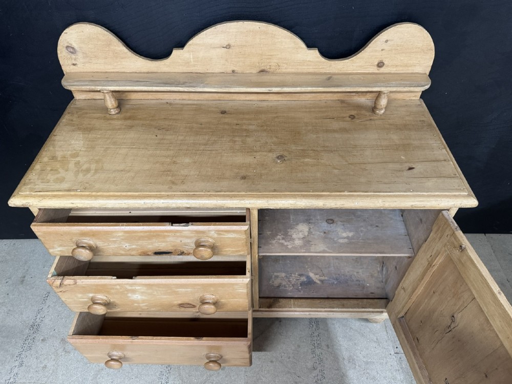 A small antique pine dresser base featuring three drawers on the left, a single panelled cupboard on the right, wooden knobs, turned bun feet, and a shaped back gallery shelf. View IV